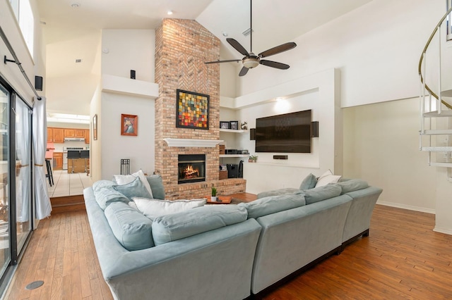 living room featuring ceiling fan, wood-type flooring, high vaulted ceiling, and a brick fireplace