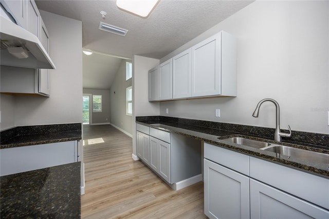 kitchen with sink, dark stone counters, lofted ceiling, white cabinets, and light wood-type flooring