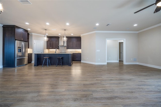 kitchen with wall chimney exhaust hood, stainless steel fridge, dark brown cabinets, and an island with sink