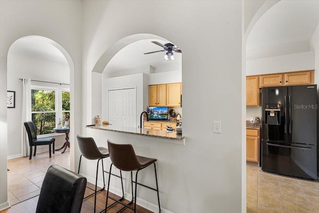 kitchen with kitchen peninsula, light brown cabinetry, black fridge, ceiling fan, and stone counters