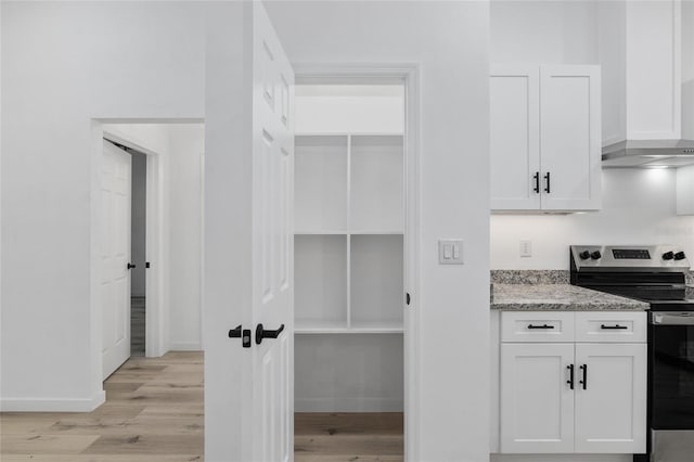 kitchen with stainless steel range with electric stovetop, white cabinets, wall chimney exhaust hood, light stone countertops, and light hardwood / wood-style floors