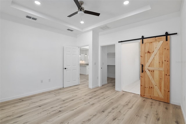 unfurnished bedroom with a raised ceiling, a barn door, a walk in closet, a closet, and light wood-type flooring