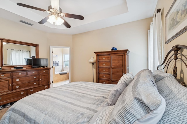 bedroom with ceiling fan, a raised ceiling, multiple windows, and ensuite bath