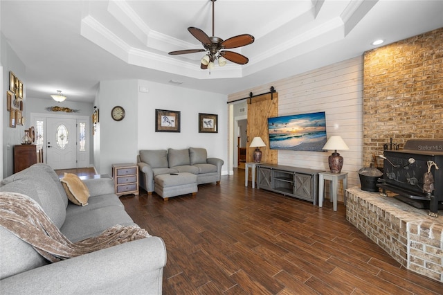 living room with dark wood-type flooring, ceiling fan, a barn door, ornamental molding, and a tray ceiling