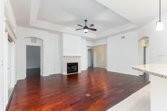 unfurnished living room with a ceiling fan, a tray ceiling, a tiled fireplace, and wood finished floors