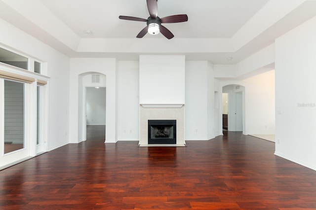 unfurnished living room with arched walkways, a raised ceiling, dark wood-type flooring, a ceiling fan, and a tile fireplace