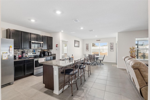 kitchen featuring a breakfast bar, backsplash, a kitchen island with sink, light stone counters, and stainless steel appliances