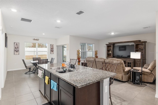 kitchen with a center island with sink, sink, light tile patterned floors, light stone counters, and dark brown cabinetry