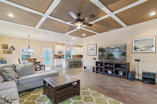 living area with ceiling fan with notable chandelier, coffered ceiling, wood finished floors, and visible vents