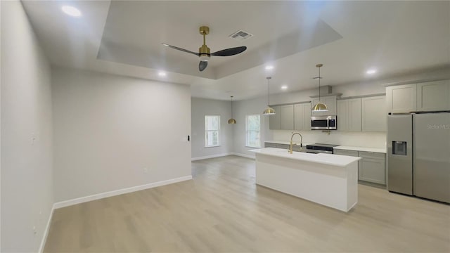 kitchen featuring appliances with stainless steel finishes, gray cabinetry, a tray ceiling, a kitchen island with sink, and ceiling fan