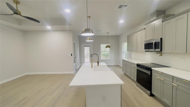 kitchen featuring decorative backsplash, gray cabinetry, stainless steel appliances, ceiling fan, and a center island with sink