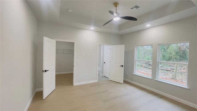 unfurnished bedroom with light wood-type flooring, a walk in closet, a tray ceiling, ceiling fan, and a closet