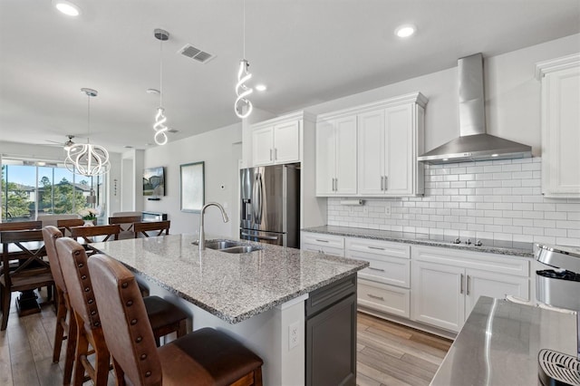 kitchen with wall chimney range hood, sink, stainless steel fridge, an island with sink, and decorative light fixtures