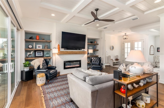 living room with coffered ceiling, wood-type flooring, a brick fireplace, built in shelves, and beamed ceiling