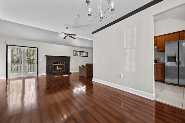 unfurnished living room with ceiling fan with notable chandelier, baseboards, vaulted ceiling, light wood-type flooring, and a brick fireplace