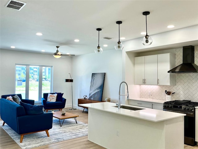 kitchen featuring gas range, sink, wall chimney range hood, a center island with sink, and hanging light fixtures