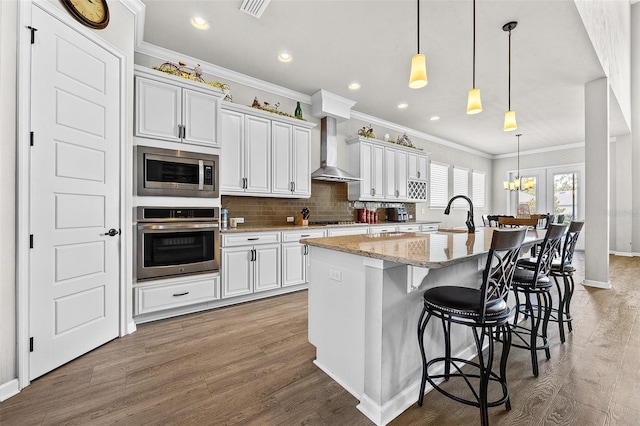 kitchen featuring a center island with sink, wall chimney exhaust hood, light stone counters, white cabinetry, and stainless steel appliances