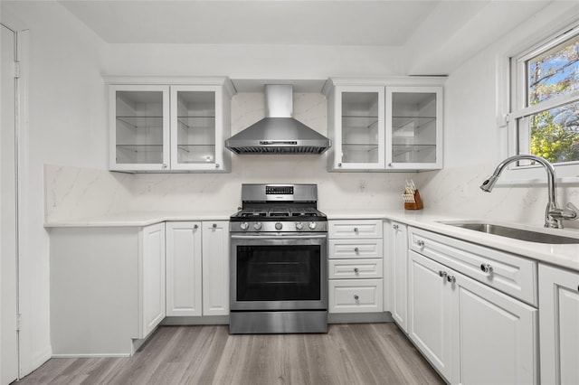 kitchen featuring glass insert cabinets, light countertops, wall chimney range hood, stainless steel range with gas cooktop, and a sink