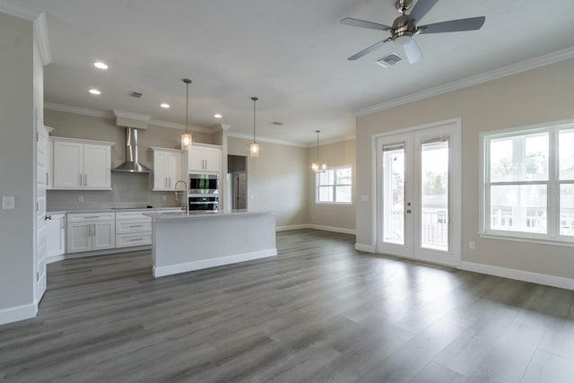 kitchen with a kitchen island with sink, white cabinets, decorative light fixtures, and wall chimney range hood