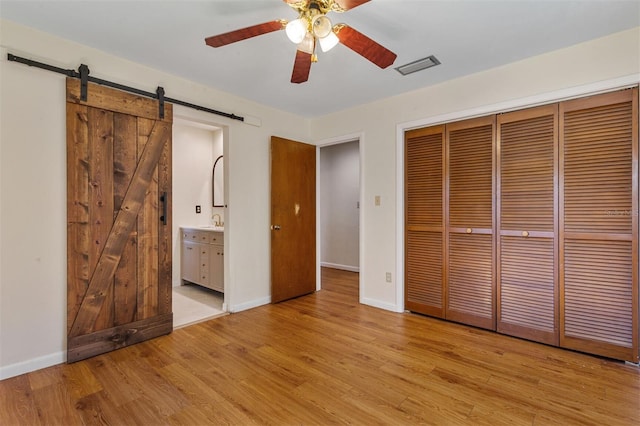 unfurnished bedroom featuring ensuite bath, ceiling fan, a barn door, light wood-type flooring, and a closet