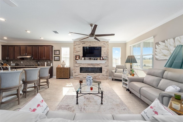 tiled living room with ceiling fan, ornamental molding, and a stone fireplace