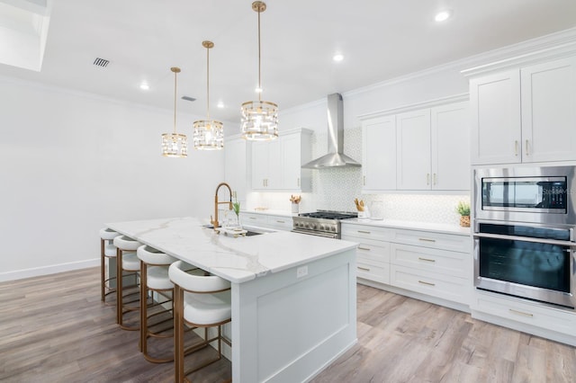 kitchen featuring white cabinets, a center island with sink, sink, wall chimney exhaust hood, and appliances with stainless steel finishes