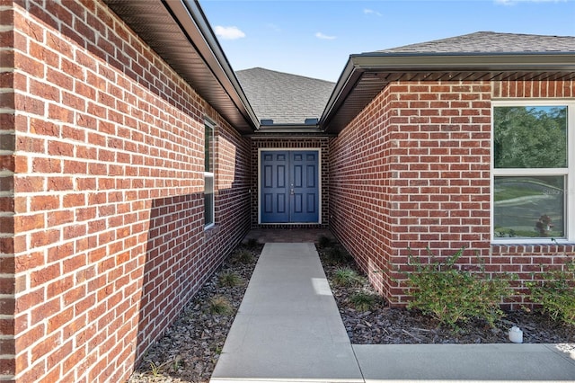 view of exterior entry with a shingled roof and brick siding