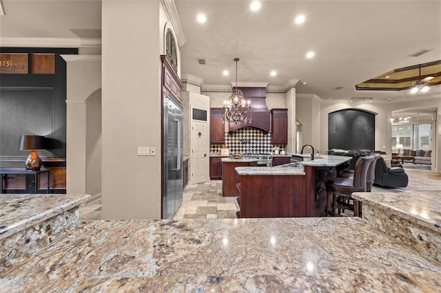 kitchen with ornamental molding, ceiling fan with notable chandelier, a breakfast bar area, premium range hood, and a center island with sink