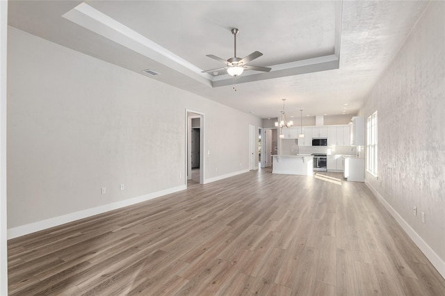 unfurnished living room with light wood-type flooring, ceiling fan with notable chandelier, a tray ceiling, and sink