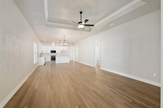 unfurnished living room featuring sink, a tray ceiling, ceiling fan with notable chandelier, and light wood-type flooring