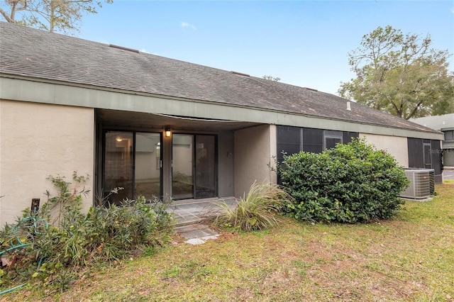 rear view of house with a shingled roof, central AC unit, a lawn, and stucco siding