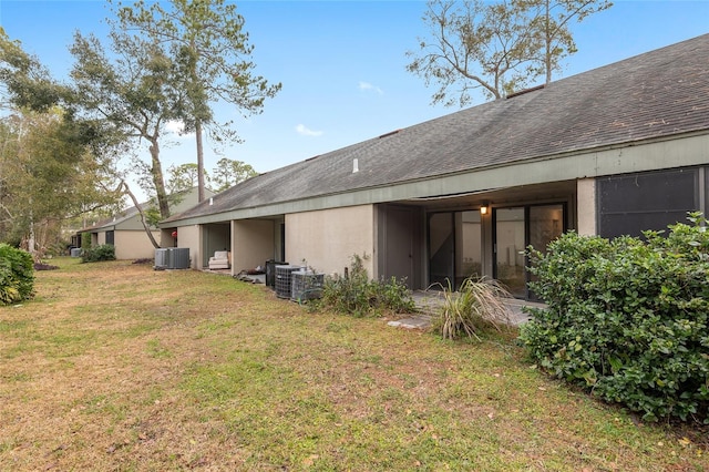 back of house featuring stucco siding, roof with shingles, central AC, and a yard