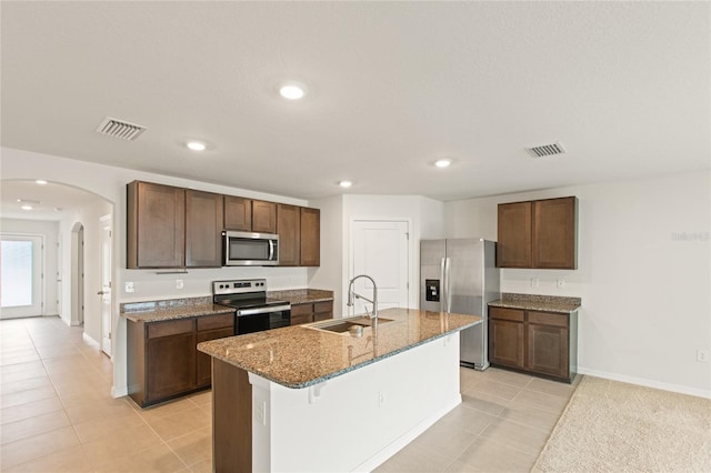 kitchen featuring sink, light tile patterned flooring, an island with sink, stainless steel appliances, and dark stone counters