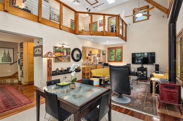 dining area with ceiling fan, hardwood / wood-style flooring, a towering ceiling, and a wood stove