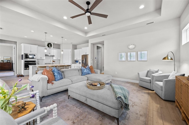 living room with light wood-type flooring, ceiling fan, a raised ceiling, and sink