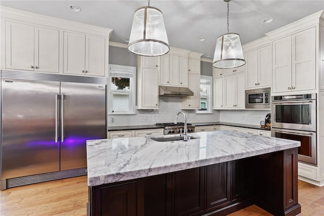 kitchen featuring sink, white cabinetry, built in appliances, an island with sink, and decorative light fixtures