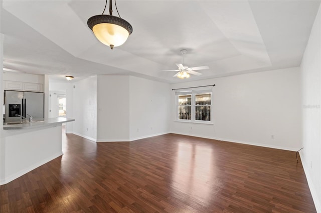 unfurnished living room featuring a raised ceiling, dark wood finished floors, baseboards, and ceiling fan