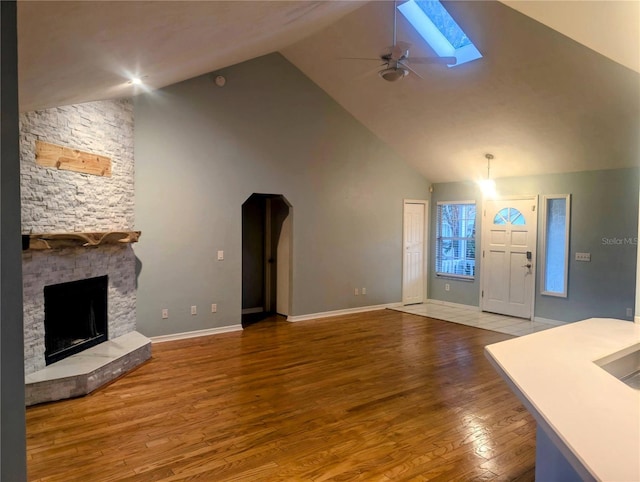 unfurnished living room with ceiling fan, a skylight, high vaulted ceiling, a fireplace, and wood-type flooring