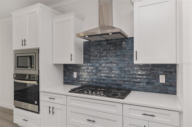 kitchen featuring wall chimney exhaust hood, crown molding, appliances with stainless steel finishes, white cabinets, and backsplash