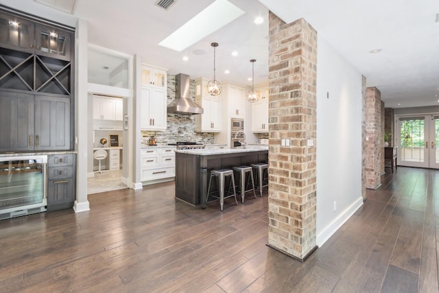 kitchen featuring white cabinets, beverage cooler, hanging light fixtures, a kitchen island with sink, and wall chimney exhaust hood