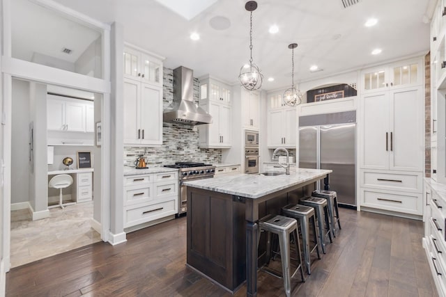 kitchen featuring sink, built in appliances, an island with sink, white cabinets, and wall chimney exhaust hood