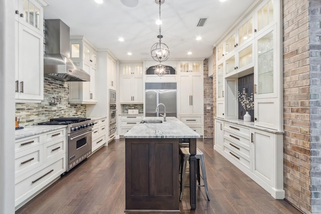 kitchen with pendant lighting, sink, built in appliances, a center island with sink, and wall chimney exhaust hood