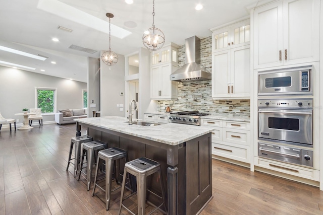 kitchen with white cabinets, appliances with stainless steel finishes, a kitchen island with sink, and wall chimney range hood