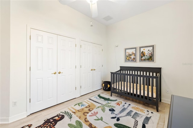 bedroom with two closets, a crib, light hardwood / wood-style floors, and ceiling fan