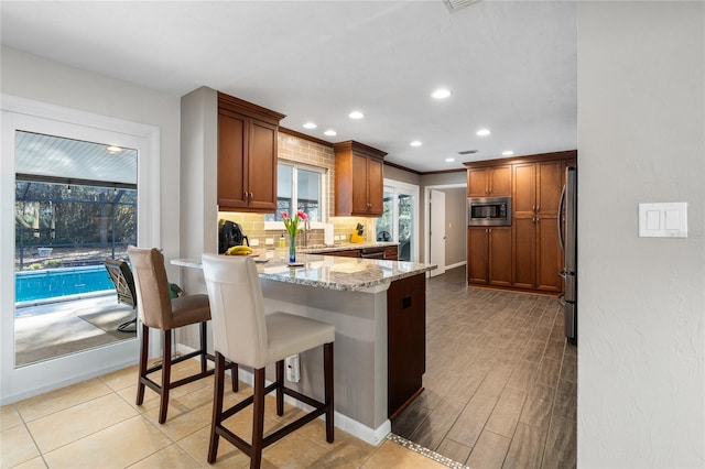 kitchen with appliances with stainless steel finishes, light stone countertops, tasteful backsplash, brown cabinetry, and a kitchen bar