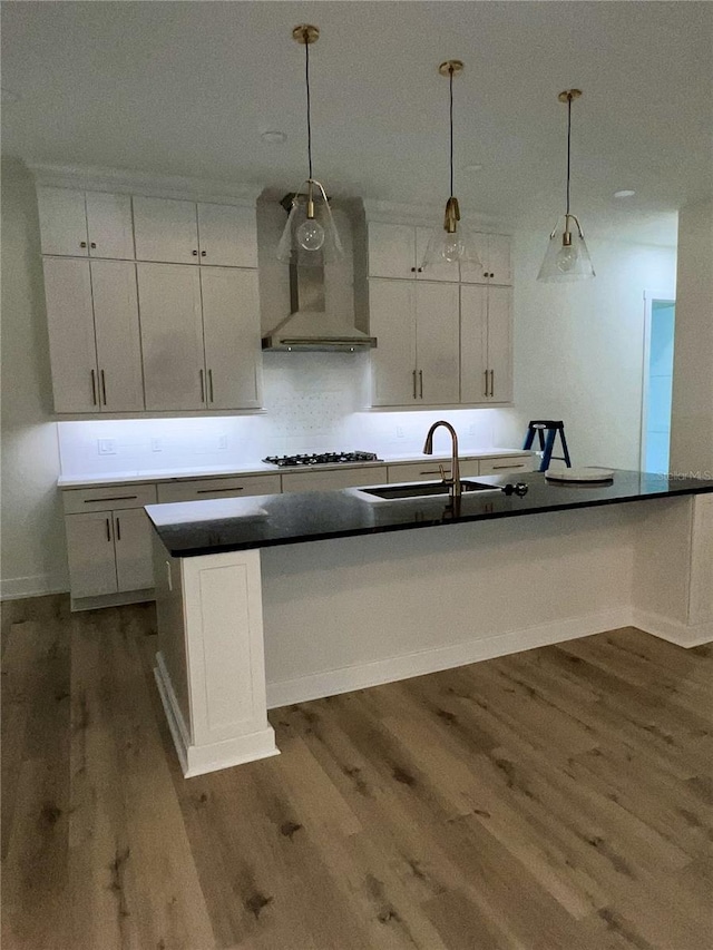 kitchen with pendant lighting, wall chimney range hood, sink, white cabinetry, and an island with sink