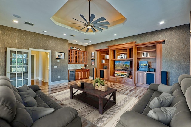 living room featuring bar, light wood-type flooring, a tray ceiling, ceiling fan, and beverage cooler