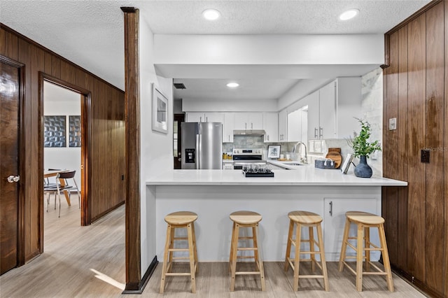 kitchen with a breakfast bar, appliances with stainless steel finishes, white cabinetry, wooden walls, and light hardwood / wood-style floors