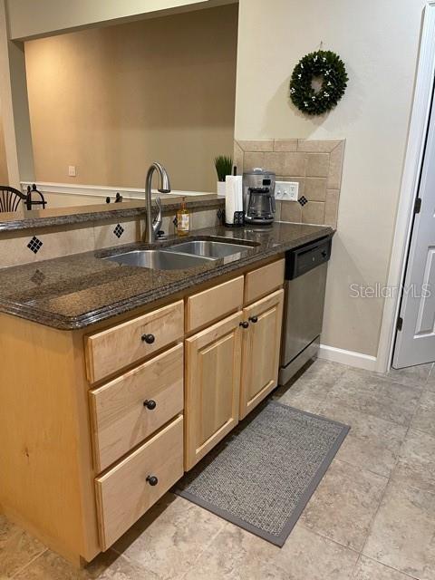 kitchen with stainless steel dishwasher, light brown cabinets, a sink, dark stone counters, and baseboards