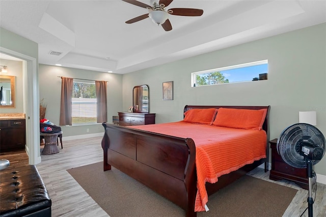 bedroom featuring connected bathroom, visible vents, baseboards, light wood-type flooring, and a tray ceiling